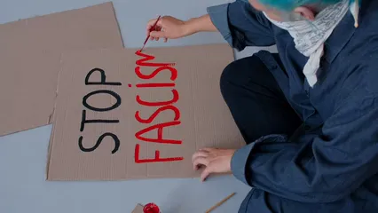 Horizontal video: A woman in a blue mask and blue hair is sitting on the ground with a sign that says stop fascism 9413320. Duration: 14 seconds. Resolution: 1920x1080